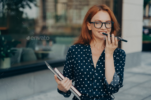 Red haired successful businesswoman records voice message keeps smartphone near mouth