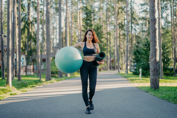 active slim woman dressed in sportswear, carries fitball and rolled up karemat