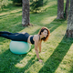 Outdoor shot of active brunette woman exercises with fitness ball poses at  green grass, dressed in active wear, enjoys sunny day and fresh air in  park, keeps fit, makes gymnastics exercises. 