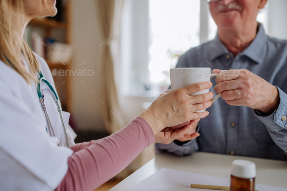 Close-up of doctor holding cup of tea and talking with senior man during medical visit at home.