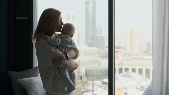 mother hugs and kisses baby standing by the window