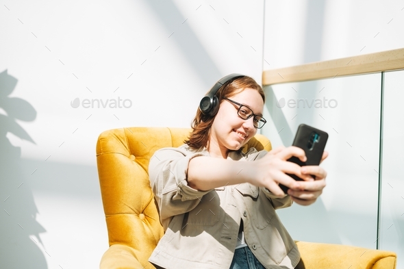 Young brunette teenager girl college student with mobile phone listen music at yellow modern chair