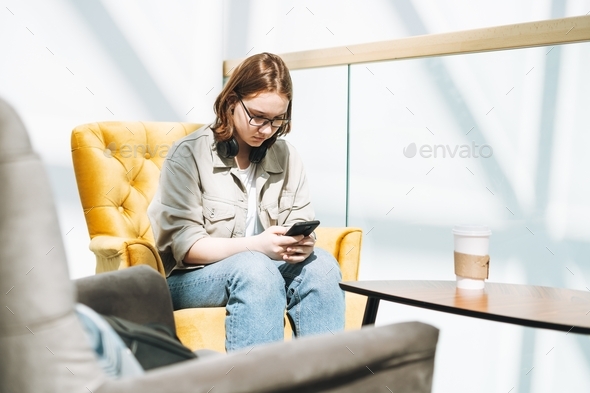 Young brunette teenager girl college student with mobile phone listen music at yellow modern chair