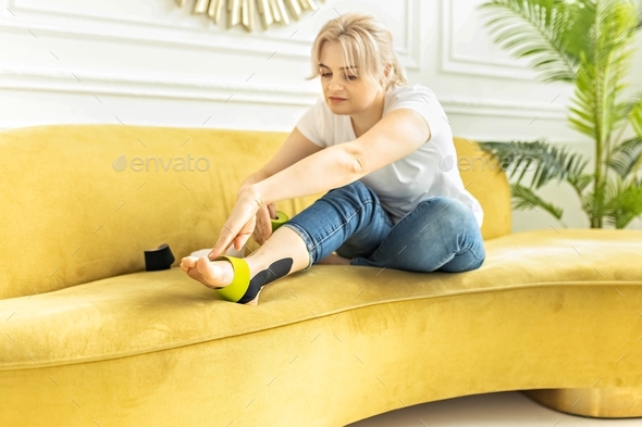 A woman is sitting on the sofa, gluing medical tape on the area of the damaged ankle joint.