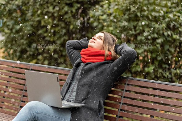 A woman, with her hands behind her head,rests after work, sits in the park a laptop on her knees.