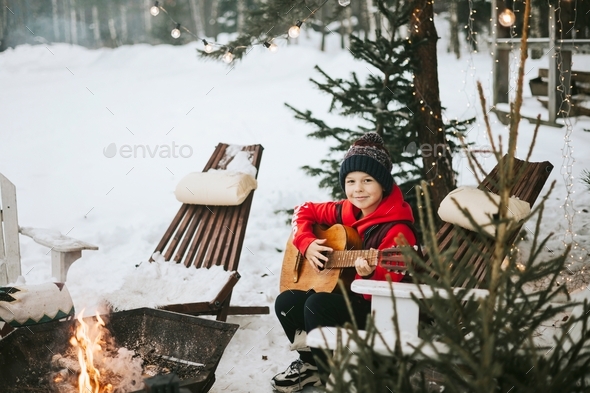 boy in a vest and a knitted hat plays the guitar outdoor near a fire pit, the concept of winter Chri
