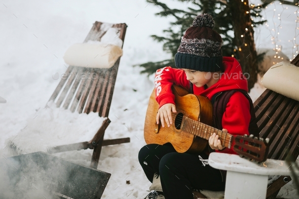 boy in a vest and a knitted hat plays the guitar outdoor near a fire pit