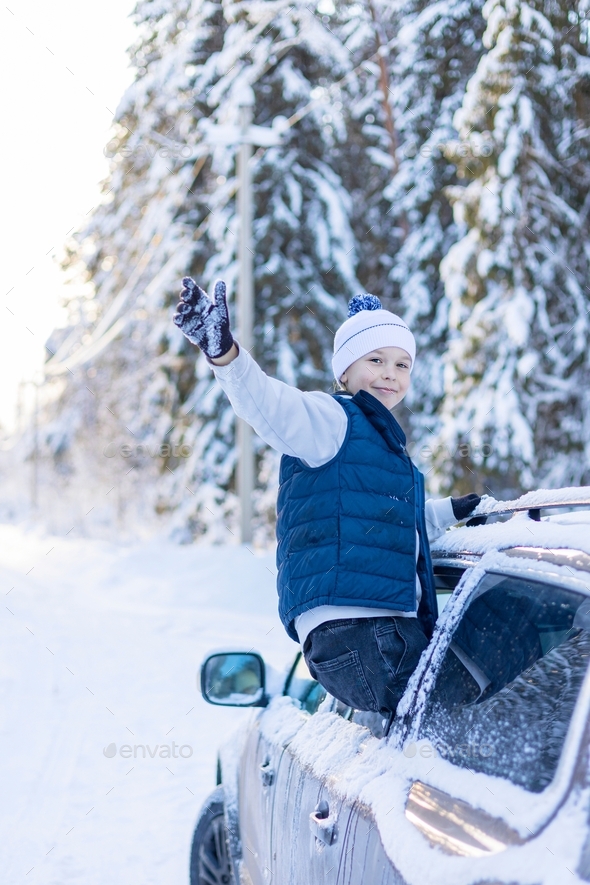teenage boy in white sweater, vest and white knitted hat in car window in snowy forest having fun, c