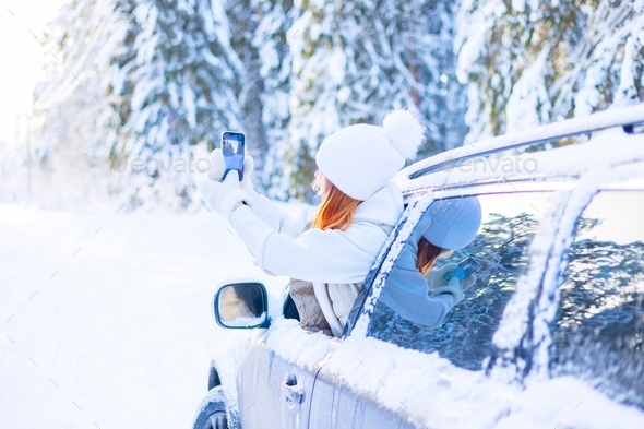 teenage girl in white sweater, vest and white knitted hat in car window in snowy forest takes selfie