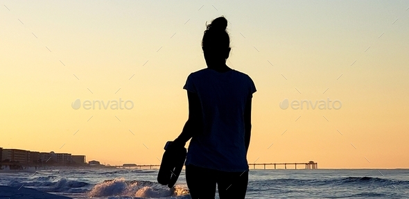 Beautiful young millennial lady carrying her flip flops while strolling down the beach at sunrise