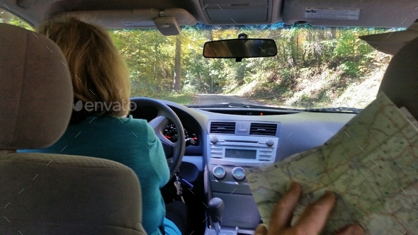 Senior woman driving car on open roads in mountains letting backseat driver guide her with map.
