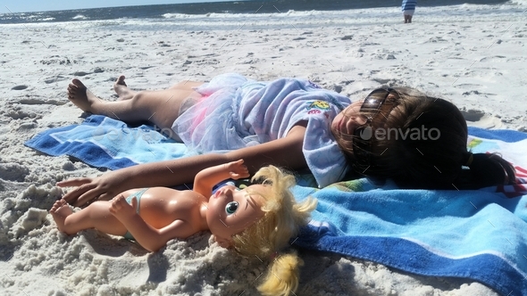Little girl sunning on the beach with her best friend Dolly but only one gets the beach towel...