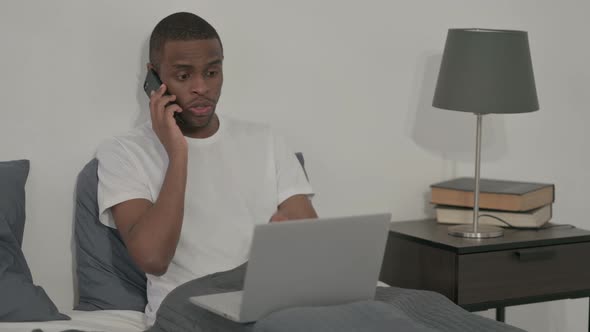 African Man with Laptop Talking on Smartphone Sitting in Bed alt