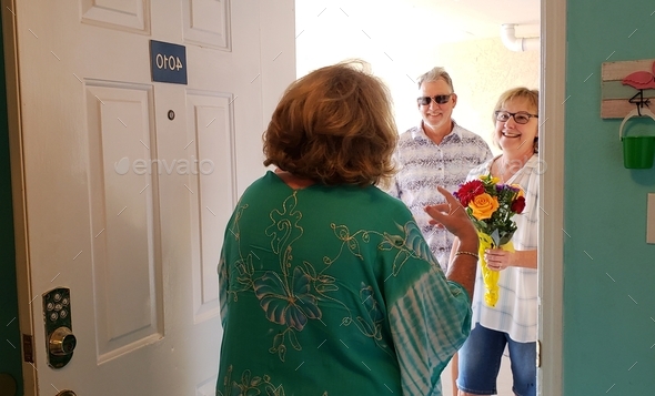 Senior woman in home opens front door to friends bringing gifts with happy faces and emotions.