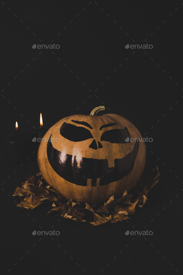 close up view of pumpkin with creepy face for halloween and dried leaves isolated on black