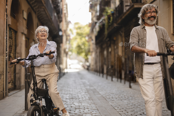 Happy mature gray hair couple in push scooter riding through the old town. Focus on elderly woman