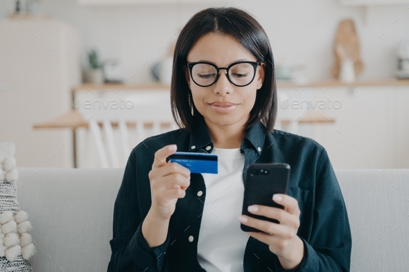 Modern female holding credit card, phone, uses mobile bank app, online banking services at home
