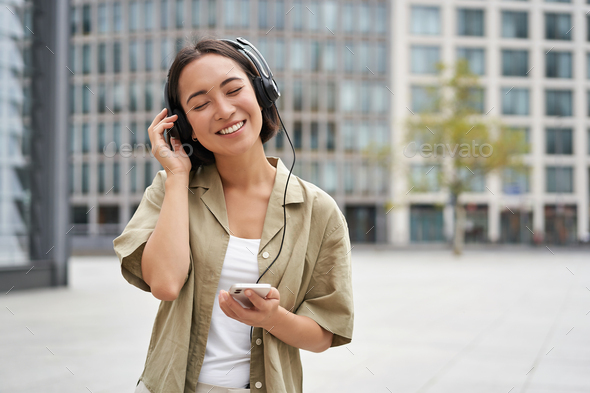 Streaming services concept. Happy asian girl listens music in headphones, holds mobile phone