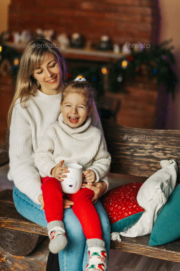 Portrait of a happy little girl sitting on her mother's lap and drinking hot chocolate on Christmas - Stock Photo - Images