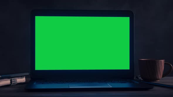 A black laptop with a green screen is on a wooden table in a dark room. alt