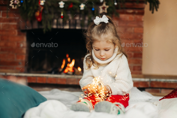 Cute little girl sitting by the fireplace with a Christmas garland.Christmas portrait, cozy style - Stock Photo - Images