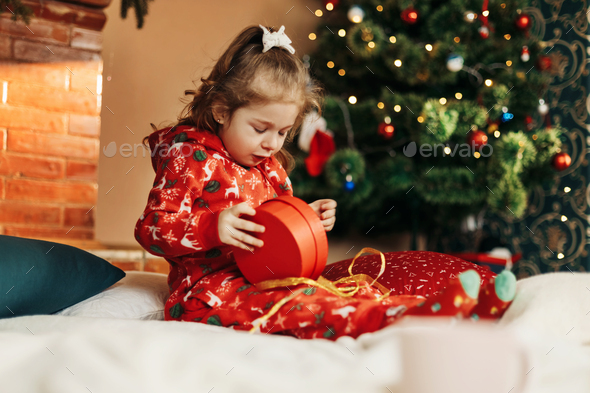 Little girl opens a Christmas present near the Christmas tree - Stock Photo - Images