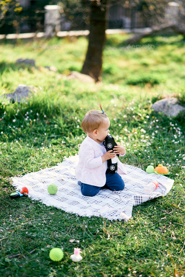Little girl sits on a blanket on the green grass and chews a toy car. Side view