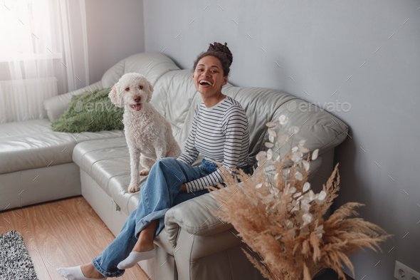 Joyful young beautiful woman owner chilling on couch with white dog poodle pet