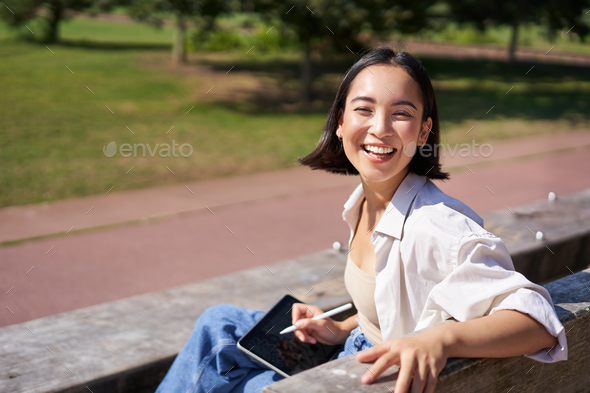 Happy korean woman sitting on bench with digital tablet and graphic pen, smiling, turning back at