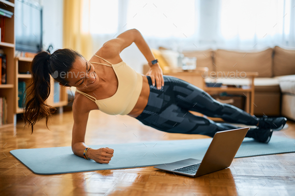 Happy woman in side plank pose having fun during online exercise class at home.