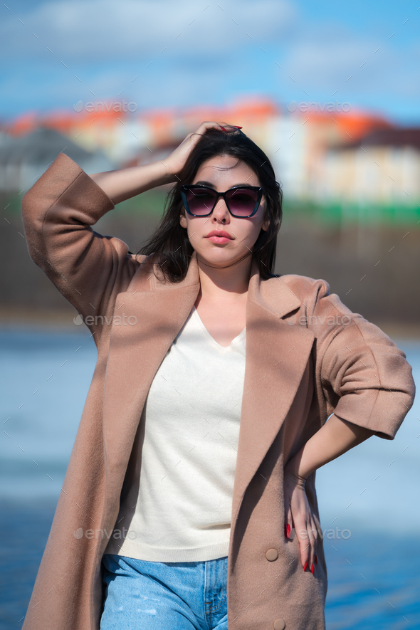 Portrait of stylish young woman with long hair, plump lips and sunglasses, dressed in beige coat - Stock Photo - Images