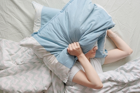 woman with dark hair in bed with a mask over her eyes falls asleep and covers her head with pillows