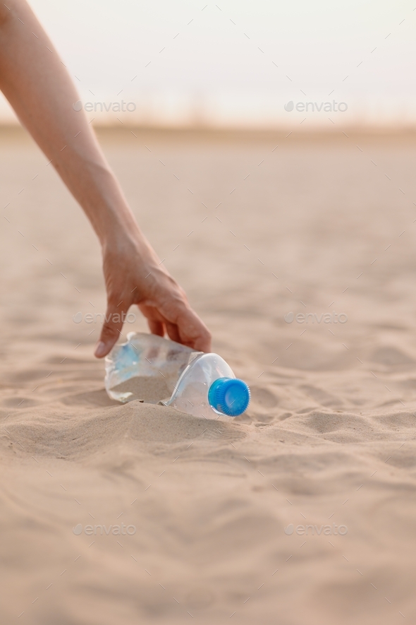 woman picks up trash and plastic bottle from beach, plastic bottle on sand on beach