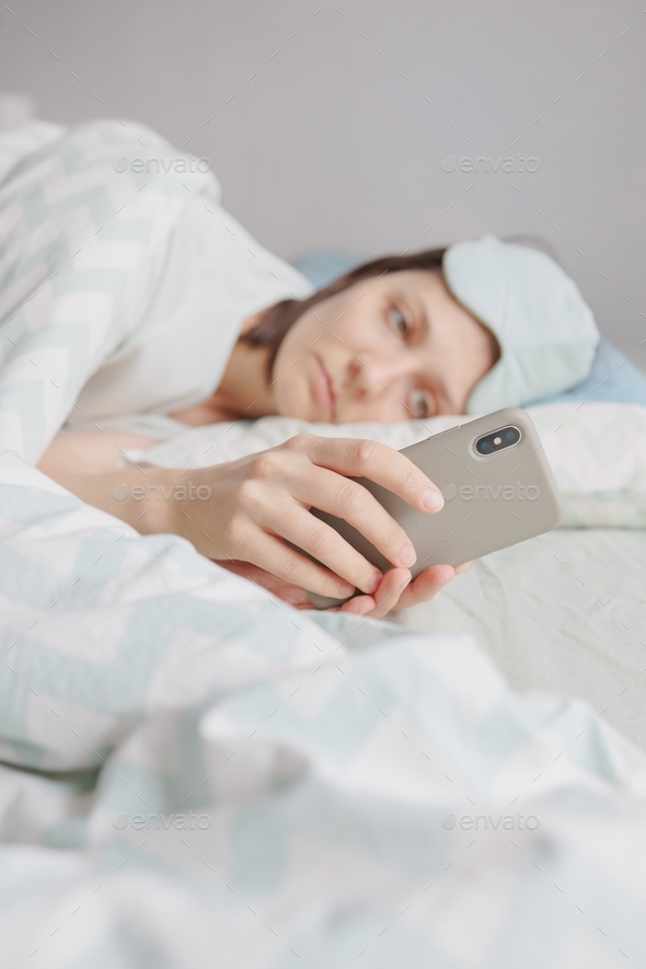 woman with dark hair in bed with a mask over her eyes falls asleep and chats on a smartphone.