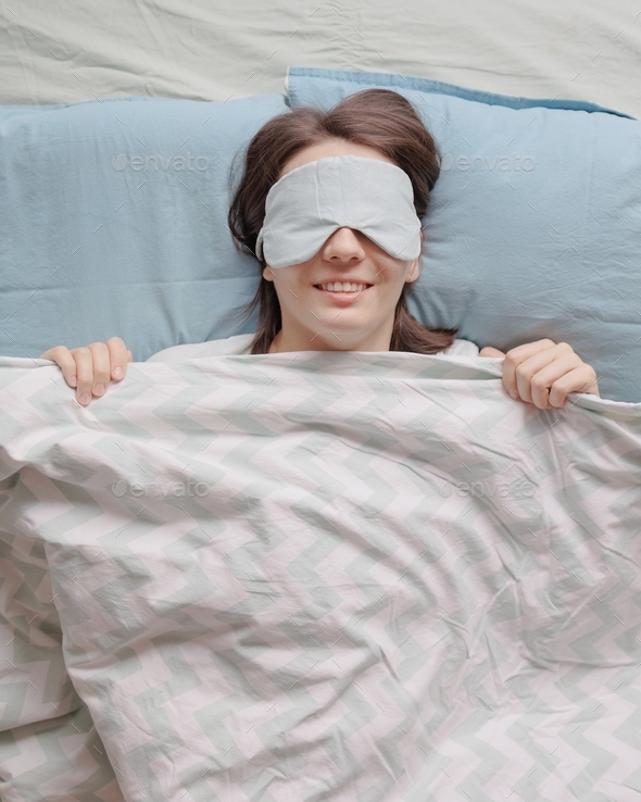  woman with dark hair in bed with a mask over her eyes falls asleep and smiles