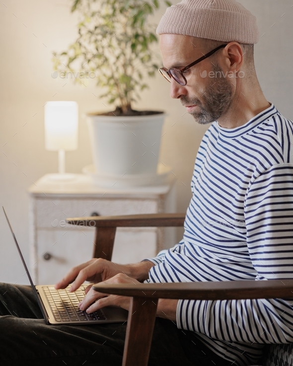 a thoughtful man with a beard is typing text on a laptop keyboard, working on a laptop in his home