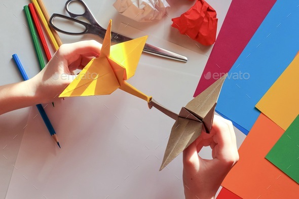 Children’s hands doing origami crane from yellow paper on white background