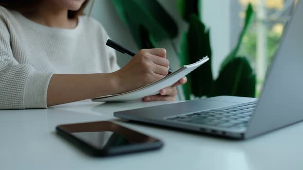 Closeup of a woman writing on a notebook with laptop computer on the table