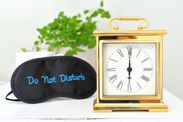 A gold clock with roman numerals setting on a dresser with a Do Not Disturb eye mask