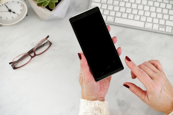 Female holding mobile phone at desk with blank black screen finger posed to click on screen mock up