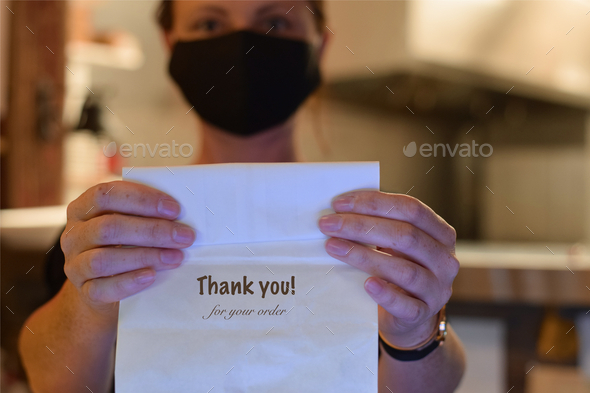 Woman restaurant employee wearing mask is holding a take out bag saying THANK YOU FOR YOUR ORDER