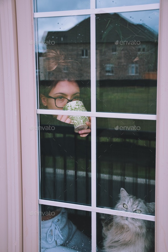 Young girl in glasses is holding a cup of tea or coffee and looking through a window on a rainy day