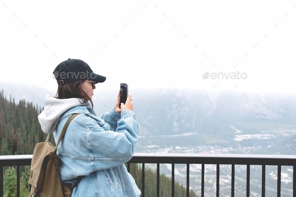 Young girl standing near railing on top of mountain taking pictures with mobile phone