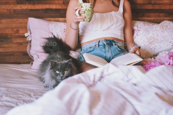 Young girl is sitting on a bed with a cup of tea or coffee reading a book with a cat by her side