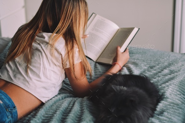 Young teenage girl laying on her bed reading a book with a cat by her side