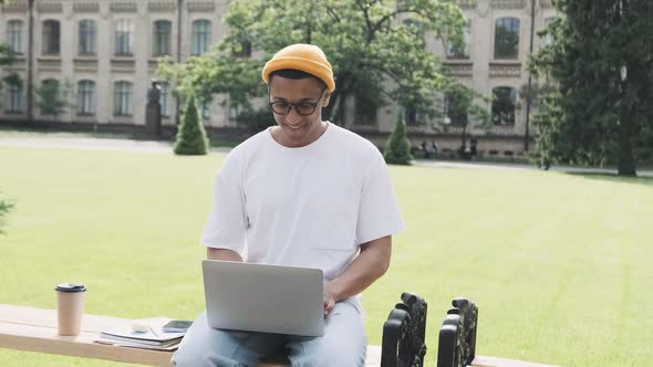 Young Cheerful Stylish African American Male Student is Sitting on a Banch Chatting on His Laptop alt
