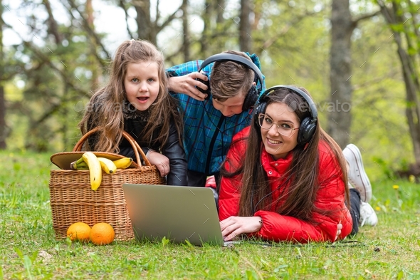 family rest on the grass next to a picnic basket in the park and watch a movie on laptop