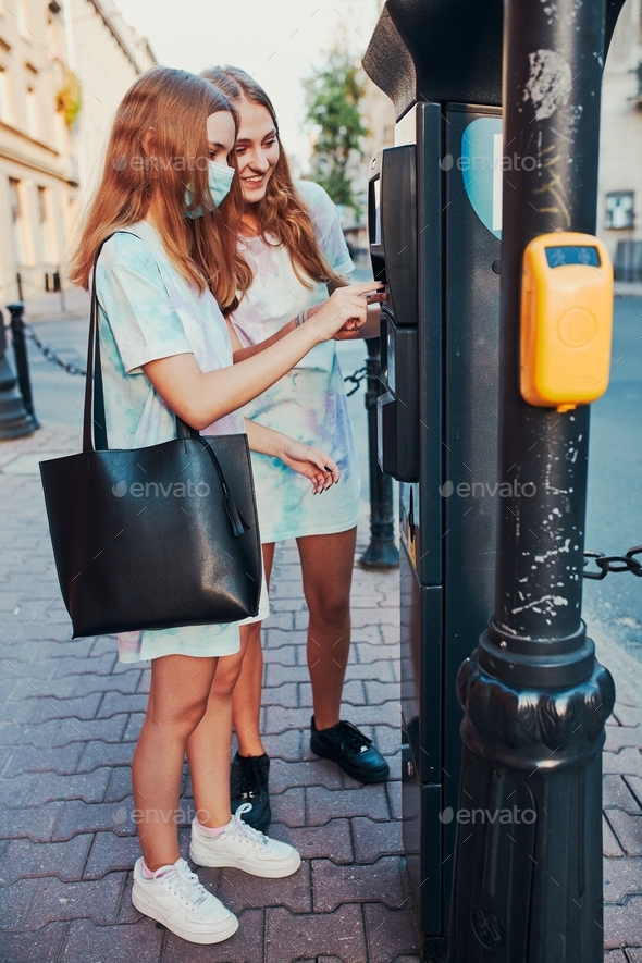 Young women girls buying parking ticket using park machine. Girl wearing face mask to avoid virus