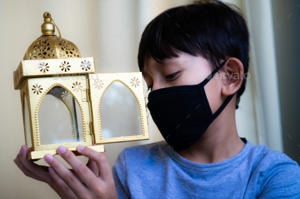 young boy wearing face mask holding islamic design lamp post while looking sad