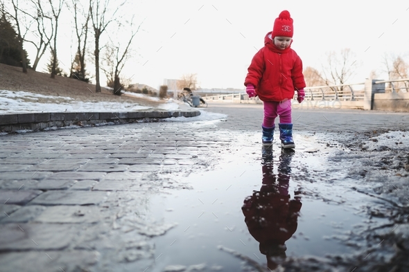 happy baby girl in boots and red jacket run and play in water puddle. funny cheerful toddler girl